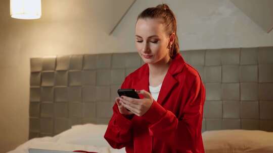 身穿红色夹克的年轻女子在床上从事智能手机