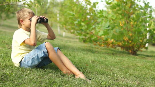 男孩坐在草地上用双筒望远镜观看