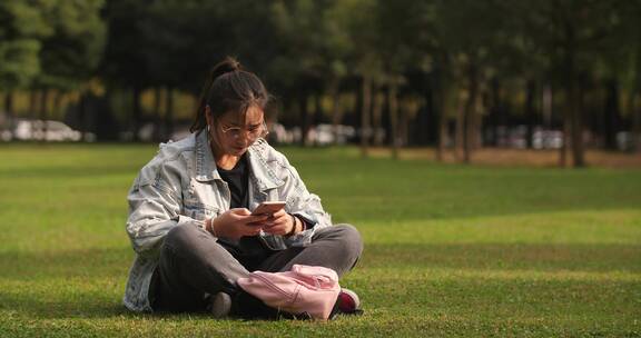 女大学生坐在校园草地上看手机浏览社交媒体