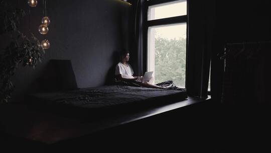 一个年轻人在卧室窗户边用笔记本电脑