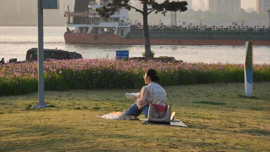 清晨阳光下一个女孩坐在江边公园草坪上看书