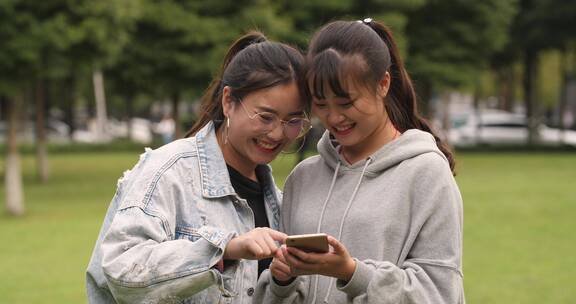 两位女大学生在校园草地上看手机网购学习