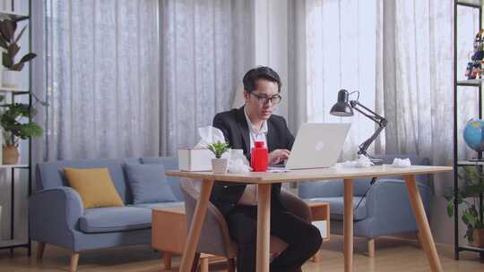 生病的亚洲商人在家客厅用笔记本电脑工作时喝热水