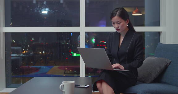 城市夜晚女商人坐在办公室窗边用电脑工作