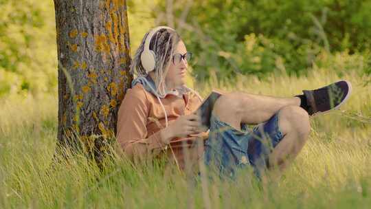 戴眼镜耳机的年轻人在夏天坐在树旁看书