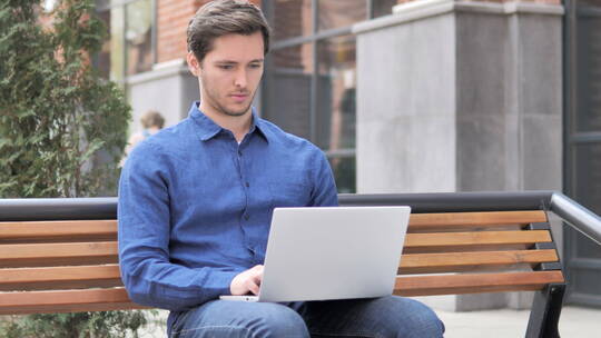 年轻男人坐在户外用笔记本电脑庆祝视频素材模板下载