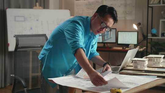 亚洲男工程师在办公室工作时拿着笔记本电脑看蓝图