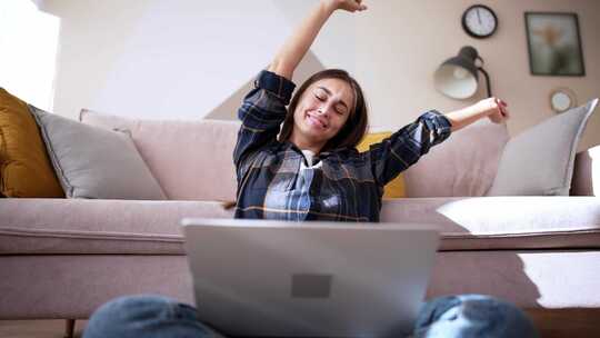 年轻女子坐在地板上的莲花位置，笔记本电脑看着屏幕，钦佩地微笑着
