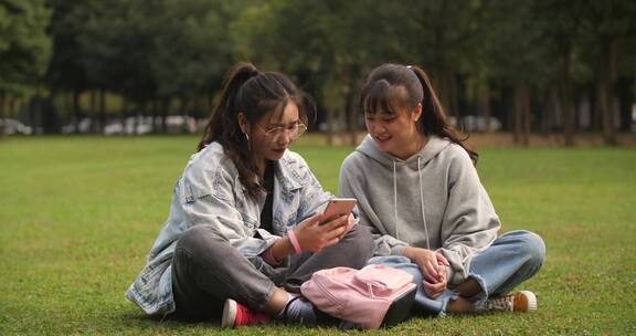 两位女大学生坐在校园草地上看手机学习交谈