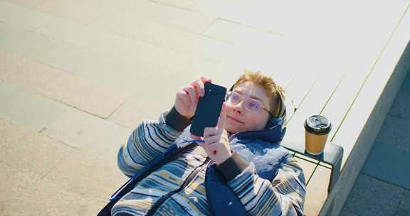 一个戴着耳机的少年男孩在网上聊天时躺在长凳上使用智能手机