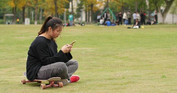 大学校园草地上女大学生坐在滑板上玩手机