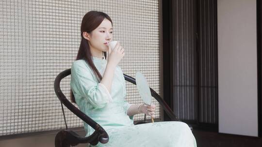 年轻旗袍女子摇着扇坐在墙边椅子喝茶休息