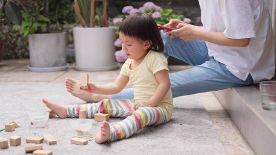 小女孩在院子里玩积木和妈妈后面看手机