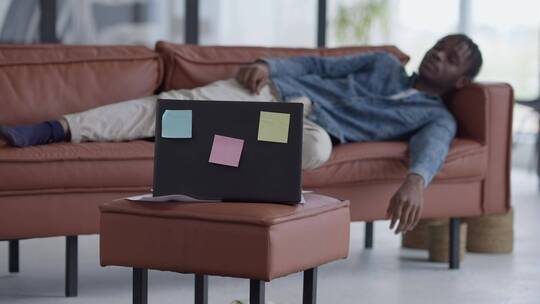 非裔年轻人睡在客厅的沙发上边上有贴着标签的笔记本视频素材模板下载