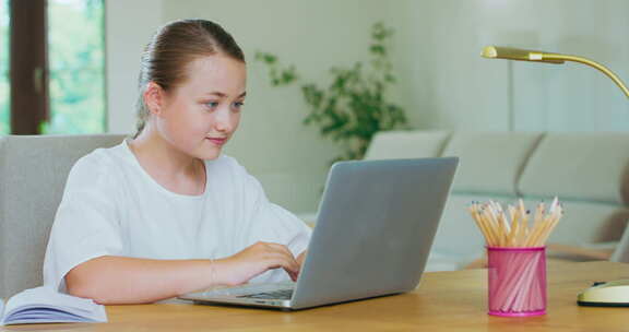 可爱的少女坐在书桌前，带着笔记本电脑，写着微笑的笔记、铅笔和一盏灯