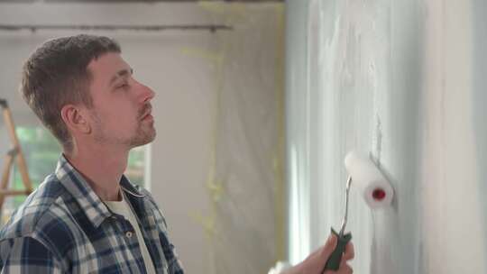 年轻人正在用油漆滚筒在窗户背景上用白色油漆粉刷墙壁