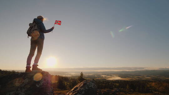 手里拿着挪威国旗的旅行者在山顶看到日出的镜头