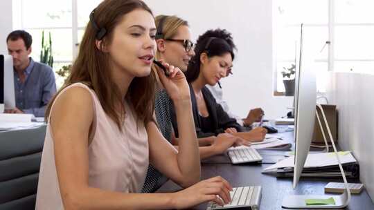 三位戴着耳机的女性在办公室的电脑前工作