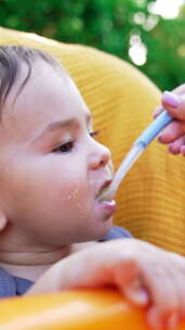 给坐在户外黄色喂食椅上的可爱幼儿喂食。