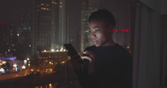 城市夜晚黑人美女在办公室窗边看手机信息
