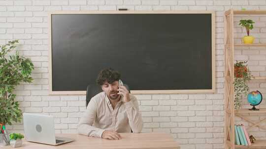 一名男教师坐在教室黑板前的书桌前，在黑板