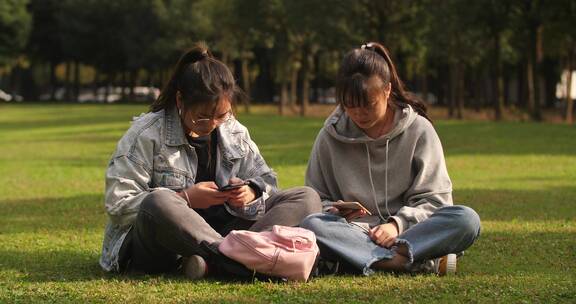 两位女大学生坐在校园草地上看手机学习交谈