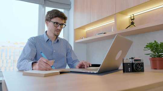 戴眼镜的年轻人在家用笔记本电脑努力工作，在笔记本上写字。
