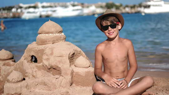 快乐晒黑的青少年男孩孩子在海滨海岸线海滩与沙城堡摆姿势享受童年