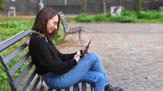 技术、设备——公园长椅上的年轻女子微笑着看着ipad上的照片视频素材模板下载