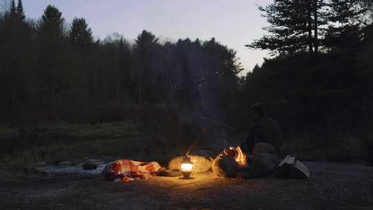 黄昏时分，一个人坐在森林里的篝火旁，手里