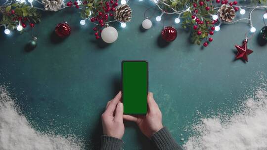 在圣诞节装饰品上方手持绿屏手机的人