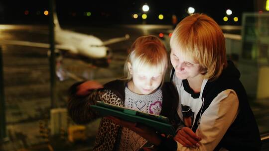 夜晚妈妈和女儿在机场的大厅里玩平板电脑