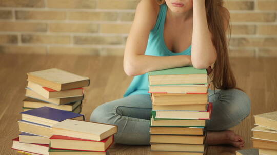 女孩看着地板上 堆叠的书籍苦笑 