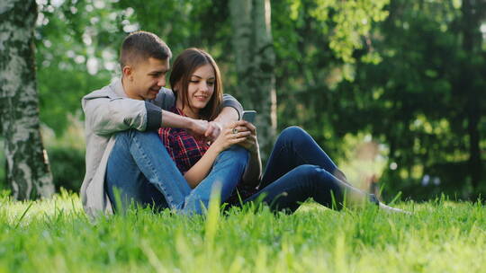 男人拥抱着女人坐在公园的草地上看着手机屏幕