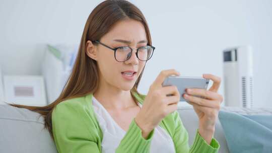 戴眼镜的亚洲女人在家用智能手机玩手机游戏。