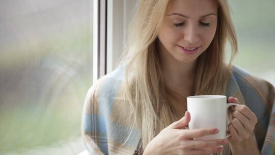 女孩披着毯子拿着一杯茶坐在窗户旁