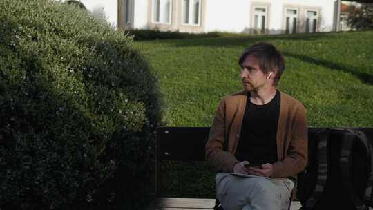 一名男子坐在公园长椅上用耳塞用智能手机通