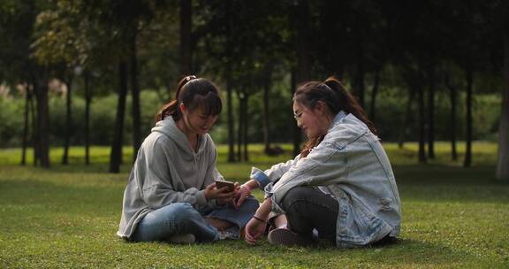 公园草地两女孩开心聊天看手机分享网购趣事