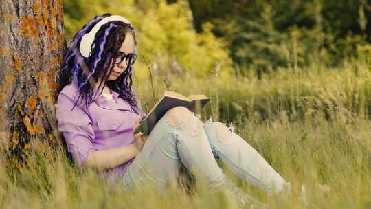 戴眼镜耳机的年轻女子在夏天坐在树旁看书