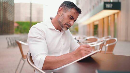 留着整齐胡子的成熟商人穿着白衬衫，在纸质记事本上写下笔记，坐在桌旁。在个人组织者中预约日常任务