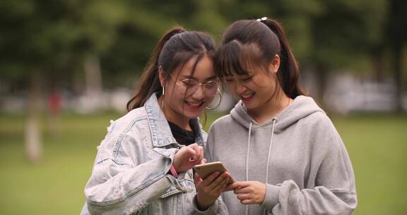 两位女大学生在校园草地上看手机网购学习