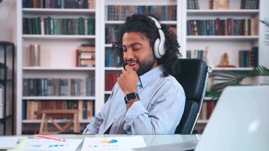 年轻的阿拉伯男子坐在办公椅上用无线耳机听音乐