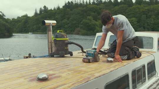 年轻人在木船前舱上用皮带打磨木板。宽镜头