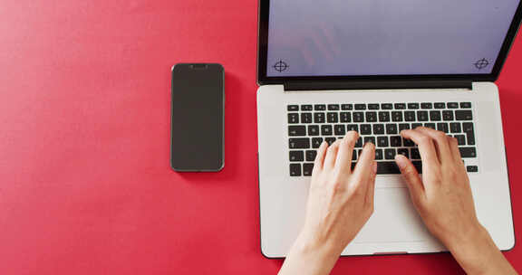 双手在带复印空间的红色桌子上使用带智能手机的笔记本电脑
