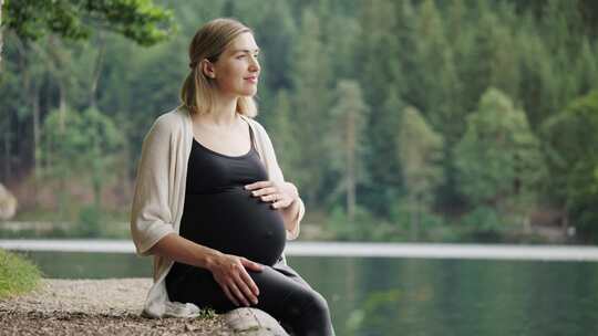 孕妇坐在山湖附近轻轻抚摸肚子