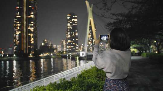 年轻女子晚上在港口用手机拍照