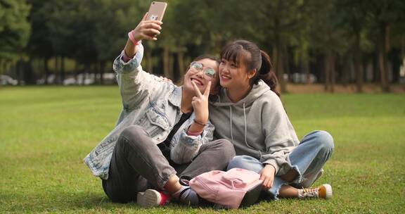 两位开心的女大学生坐在校园草坪上手机自拍