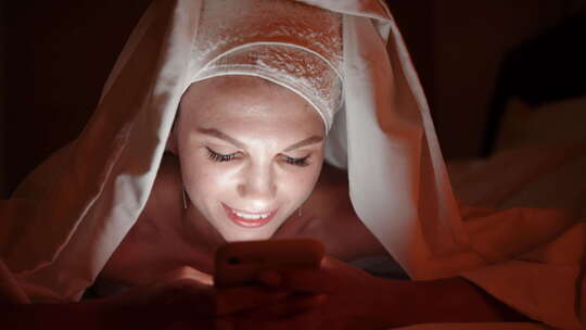 睡前在卧室毯子下使用智能手机的美丽年轻女子视频素材模板下载