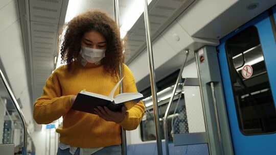 手持年轻的深色皮肤女性乘坐地铁，戴着医用口罩读一本书