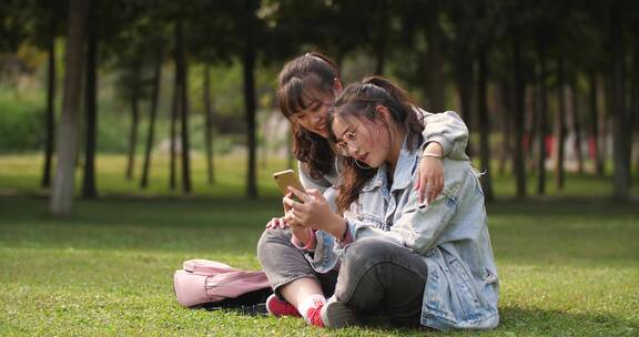 两位女大学生坐在校园草地上看手机网购学习